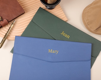 May include: A blue and green leather document holder with the names "Mary" and "Jean" in gold lettering. A brown leather journal, a gold pen, and a tan hat are also visible.