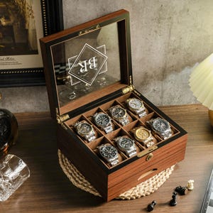 May include: A dark wood watch box with a glass lid featuring a white geometric logo with the letters BR. The interior is divided into eight compartments, each containing a silver-toned wristwatch with a metal link band and a dark watch face.