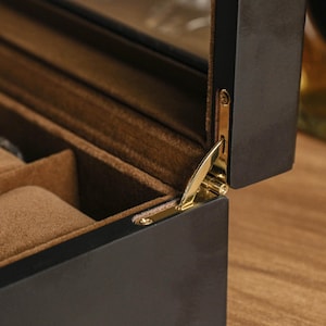 May include: A partially open, dark wood jewelry box with a gold-colored hinge. The interior is lined with a soft, brown fabric and features compartments for storing jewelry. The box is on a wooden surface.
