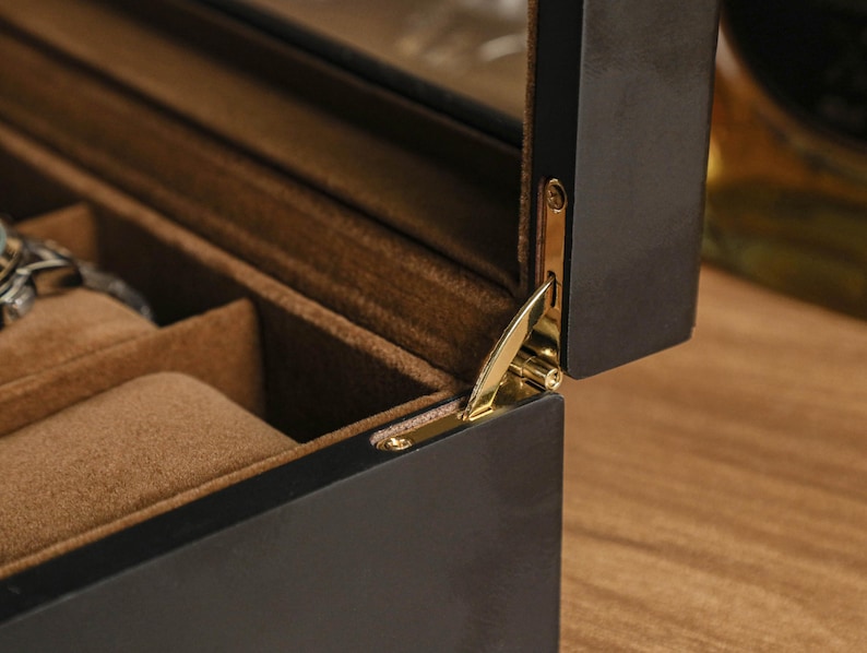 May include: A partially open, dark wood jewelry box with a gold-colored hinge. The interior is lined with a soft, brown fabric and features compartments for storing jewelry. The box is on a wooden surface.