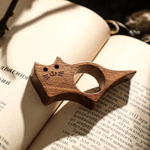 May include: A wooden cat-shaped thumb page holder, resting on an open book. The page holder is made of dark wood and features a carved cat face. The book has text in a foreign language.