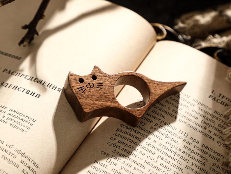 May include: A wooden cat-shaped thumb page holder, resting on an open book. The page holder is made of dark wood and features a carved cat face. The book has text in a foreign language.