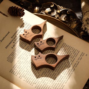 May include: Three wooden cat-shaped book page holders. Each has a circular opening and a carved cat face. The holders are made of a dark wood and rest on the open pages of a book.