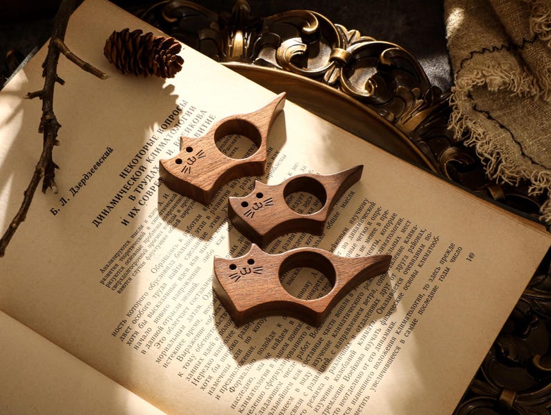 May include: Three wooden cat-shaped book page holders. Each has a circular opening and a carved cat face. The holders are made of a dark wood and rest on the open pages of a book.