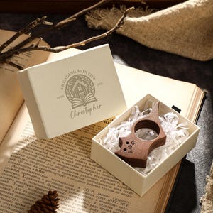 May include: A wooden cat-shaped book page holder rests in a white box with shredded paper. The box sits on an open book next to a box with the text "READING MONTH" and the name "Christopher".