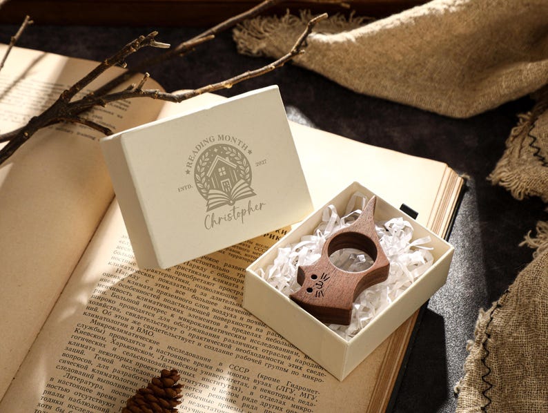 May include: A wooden cat-shaped book page holder rests in a white box with shredded paper. The box sits on an open book next to a box with the text "READING MONTH" and the name "Christopher".