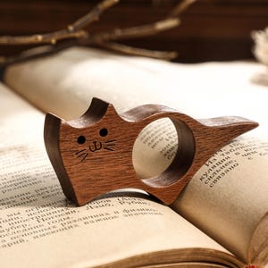May include: A wooden cat-shaped page holder rests on an open book. The page holder is made of dark wood and features a carved cat face with a smiling mouth and whiskers. The book has aged pages and is open to a page of text.