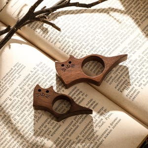 May include: Two wooden cat-shaped book holders rest on an open book. The cat-shaped holders are brown with carved details, including eyes, noses, and whiskers. The book has aged pages and text.
