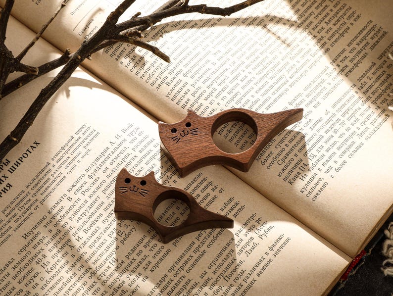 May include: Two wooden cat-shaped book holders rest on an open book. The cat-shaped holders are brown with carved details, including eyes, noses, and whiskers. The book has aged pages and text.