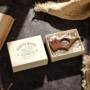 May include: A wooden cat-shaped book page holder rests in a cream-colored box filled with shredded paper. The box is open next to a cream-colored square box with the text "READING MONTH" and a floral design. An open book is in the background.