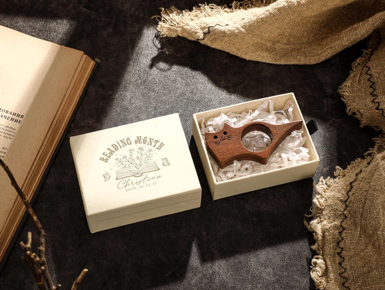 May include: A wooden cat-shaped book page holder rests in a cream-colored box filled with shredded paper. The box is open next to a cream-colored square box with the text "READING MONTH" and a floral design. An open book is in the background.
