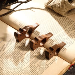 May include: Four wooden book page holders, each with a unique design, rest on the open pages of a vintage book. The holders are a rich brown color, and the book's pages are aged.