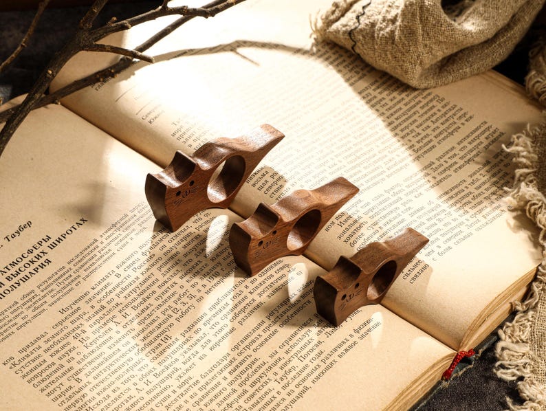 May include: Four wooden book page holders, each with a unique design, rest on the open pages of a vintage book. The holders are a rich brown color, and the book's pages are aged.