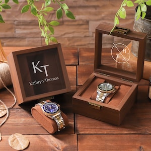 May include: Two wooden watch display boxes, one open and one closed, with a watch in each. The closed box has the initials "KT" and the name "Kathryn Thomas". The open box displays a watch with a silver and gold band. The background includes greenery and a candle.