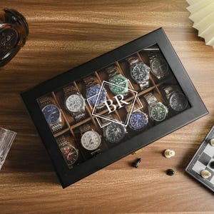May include: A black watch display case with a glass top, filled with various wristwatches. The watches have different colored faces and bands. The initials "BR" are superimposed on the watches. A chess board and a glass are also visible.