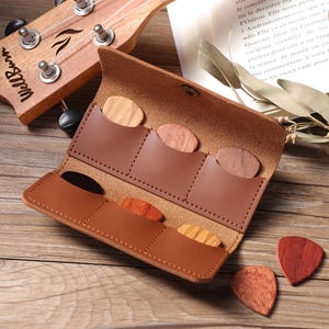 May include: A brown leather guitar pick holder, open to display several wooden picks. A wooden ukulele is visible in the background. The holder has pockets for the picks, which are made from different types of wood. The picks are various colors.