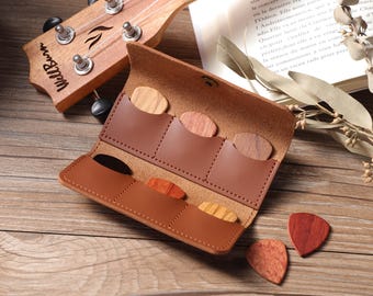 May include: Open brown leather guitar pick holder with several wooden picks of varying colors. A wooden ukulele is in the background. The holder is open, revealing pockets for the picks. The picks are made of different types of wood.