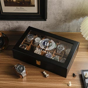 May include: A black wooden watch display case with a glass top, holding several wristwatches. The case is personalized with the name "Wilson" in a circular emblem. A wristwatch rests on a brown stand nearby. A bottle of liquor and a chess set are also visible.