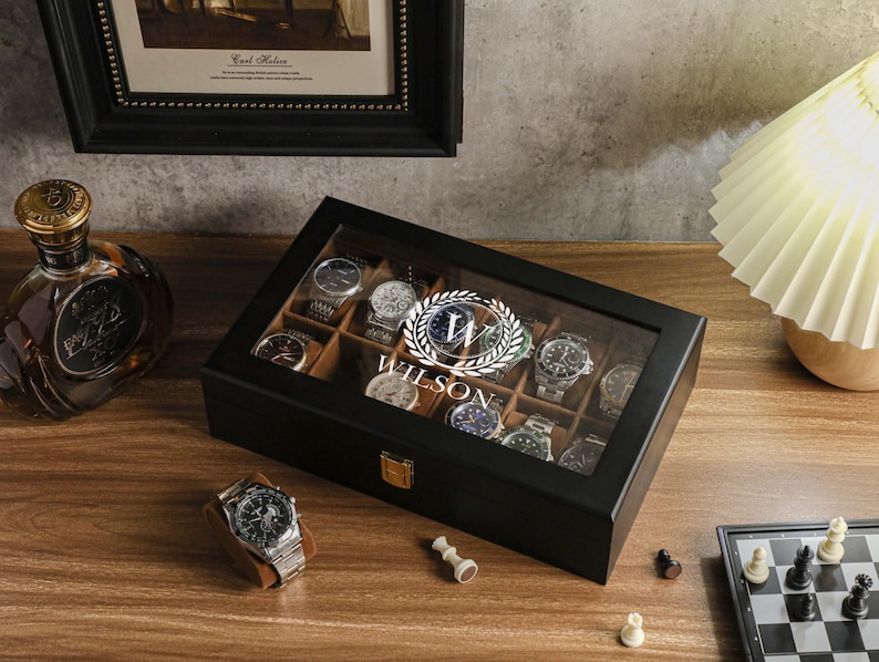 May include: A black wooden watch display case with a glass top, holding several wristwatches. The case is personalized with the name "Wilson" in a circular emblem. A wristwatch rests on a brown stand nearby. A bottle of liquor and a chess set are also visible.