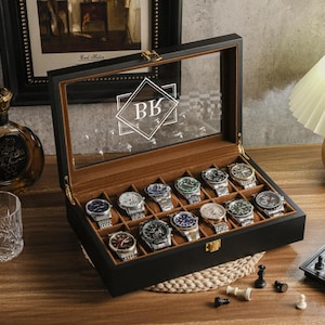 May include: A black watch display case with a glass top, holding 15 assorted wristwatches. The case is open, revealing a brown interior. The initials "BK" are visible on the glass. A chess set and a bottle of liquor are in the background.