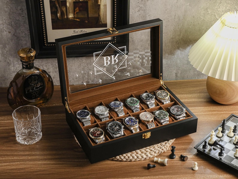 May include: A black watch display case with a glass top, holding 15 assorted wristwatches. The case is open, revealing a brown interior. The initials "BK" are visible on the glass. A chess set and a bottle of liquor are in the background.
