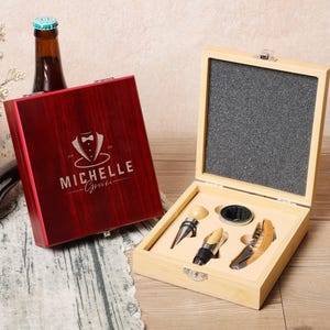 Puede incluir: Una caja de madera con una tapa roja grabada con "Michelle Gifts" y un gráfico de esmoquin. La caja contiene un juego de abridor de botellas con un tapón de botella, un cortador de papel de aluminio y un sacacorchos.