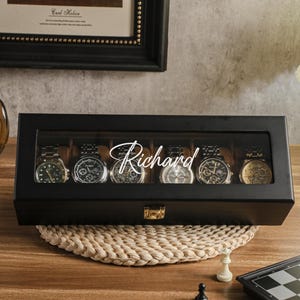 May include: A black watch display case with a glass top, holding six wristwatches. The name "Richard" is written across the glass. The case sits on a woven mat, with a chess set in the foreground.