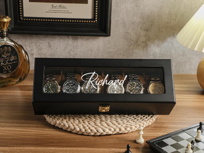 May include: A black watch display case with a glass top, holding six wristwatches. The name "Richard" is written across the glass. The case sits on a woven mat, with a chess set in the foreground.