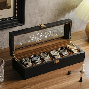 May include: A black watch display case with a clear glass top, holding six wristwatches. The interior is lined with brown fabric. The glass top has the letters "V 2 W" etched on it. A chess set and a glass are on the table.