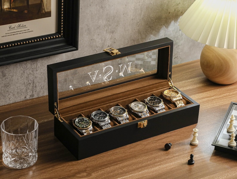 May include: A black watch display case with a clear glass top, holding six wristwatches. The interior is lined with brown fabric. The glass top has the letters "V 2 W" etched on it. A chess set and a glass are on the table.