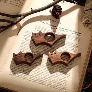 May include: Three wooden cat-shaped book holders. Each holder is carved from dark wood and features a cat face design. They are resting on the open pages of a book, with text visible.