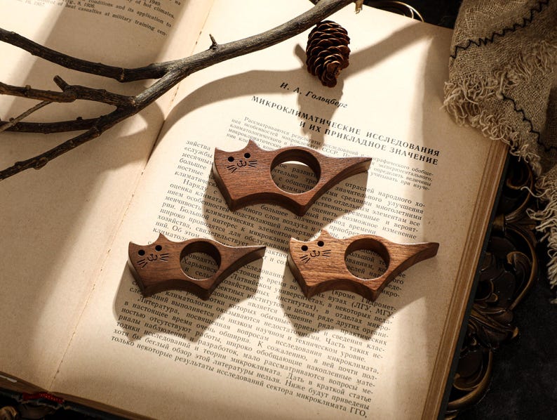 May include: Three wooden cat-shaped book holders. Each holder is carved from dark wood and features a cat face design. They are resting on the open pages of a book, with text visible.