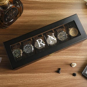 May include: A black watch display case with six watches, each with a different face design. The case has a clear top and the initials "RN" are displayed. A bottle of liquor and a chess set are also visible.