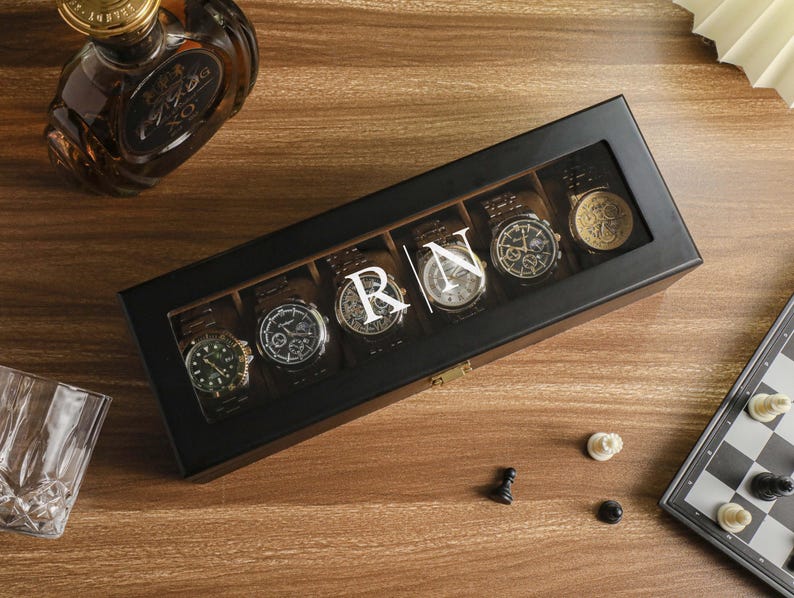 May include: A black watch display case with six watches, each with a different face design. The case has a clear top and the initials "RN" are displayed. A bottle of liquor and a chess set are also visible.