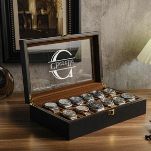 May include: A black wooden watch display case with a clear glass top, showcasing multiple wristwatches. The interior is lined with a brown fabric. The glass top features the name "Guelle" in a white, stylized font. The case is open, revealing the watches.