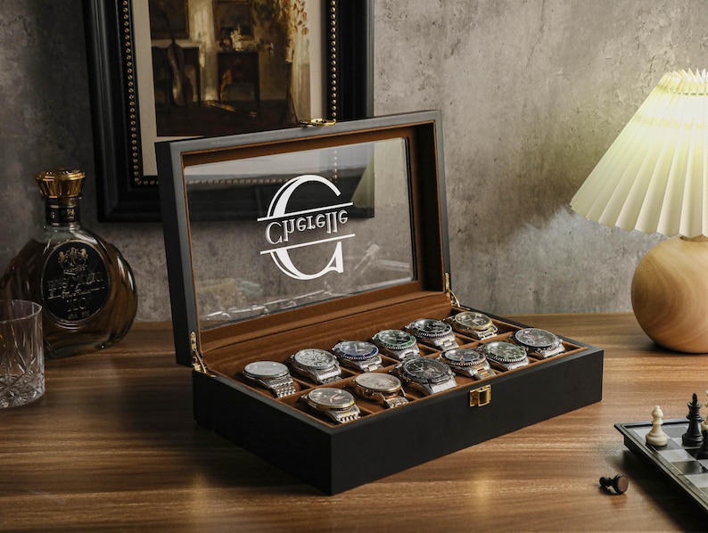 May include: A black wooden watch display case with a clear glass top, showcasing multiple wristwatches. The interior is lined with a brown fabric. The glass top features the name "Guelle" in a white, stylized font. The case is open, revealing the watches.