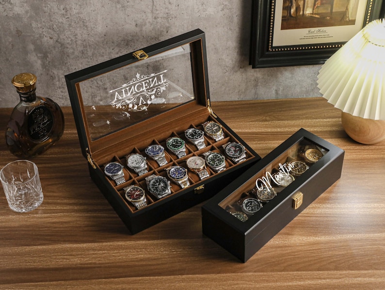 May include: Two black wooden watch display cases with glass lids. One case is open, revealing multiple watches. The other case is closed, with the name "Martin" in script on the lid. A bottle of liquor and a glass are in the background.