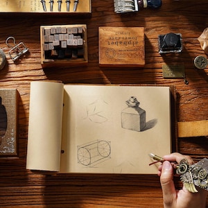 May include: A brown leather bound journal with blank pages. The journal is open to a page with pencil sketches of a bottle and a cylinder. The journal is on a wooden surface with other craft supplies.