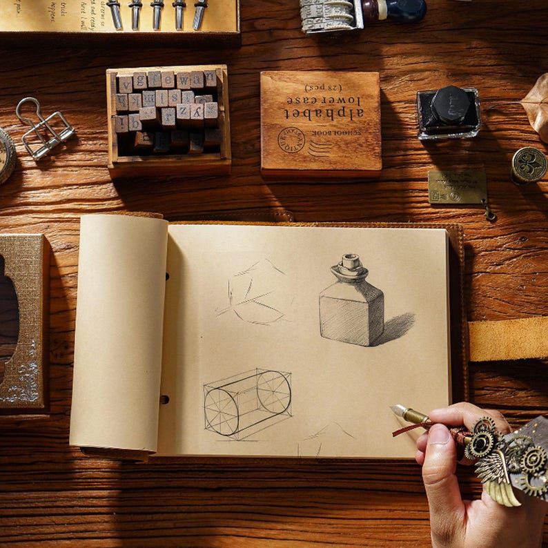 May include: A brown leather bound journal with blank pages. The journal is open to a page with pencil sketches of a bottle and a cylinder. The journal is on a wooden surface with other craft supplies.