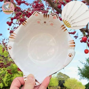 Peut inclure: Bol en céramique blanche en forme de chat avec des rayures brunes et des empreintes de pattes. Le bol est légèrement incliné et tenu dans une main. L'arrière-plan est une image floue d'un arbre avec des baies rouges.