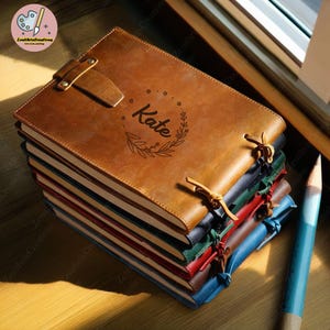 May include: A stack of brown leather journals with a personalized inscription that reads "Kate". The top journal has a leather strap and a decorative closure.