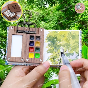 Op de afbeelding: Een houten aquarelpalette met een magnetische sluiting, een klein schetsboek en een transparante plastic penseelpen. Het palet heeft 9 aquarelverfpannetjes en een lege ruimte om kleuren te mengen. Het schetsboek is open op een pagina met een aquarel schilderij van bomen.