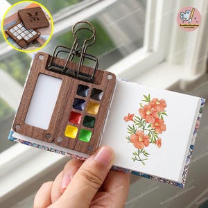 Puede incluir: Una paleta de acuarela de madera con un cuaderno de bocetos integrado. La paleta tiene 12 pastillas de acuarela y un peque&ntilde;o espacio para un pincel. El cuaderno de bocetos tiene una ilustraci&oacute;n floral en la p&aacute;gina.