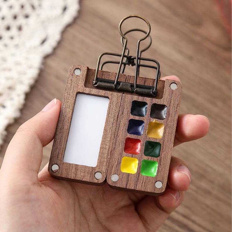 Op de afbeelding: Een houten aquarelpalette met een ingebouwde clip. De palette heeft een kleine, rechthoekige ruimte voor een penseel en negen verfputjes gevuld met kleurrijke aquarelverf.