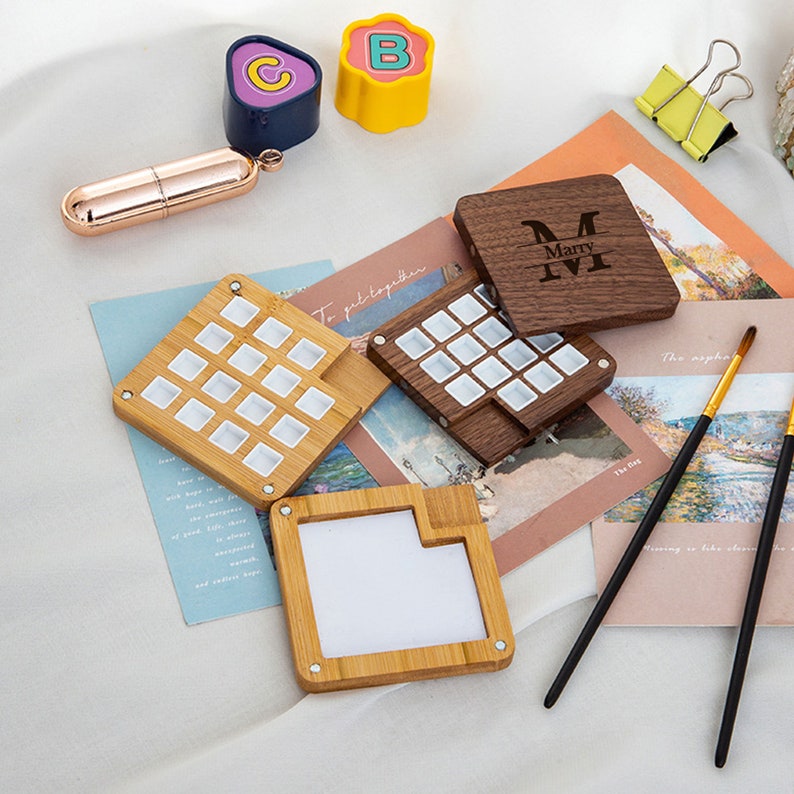 Op de afbeelding: Een set van drie houten aquarelpaletten. Twee paletten zijn gemaakt van lichtbruin hout en hebben elk 18 vakjes. De derde palette is gemaakt van donkerbruin hout en heeft een enkel compartiment met de naam "Marry M" gegraveerd op de deksel.