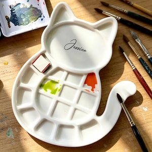 May include: A white ceramic cat-shaped paint palette with the name "Jessica" in cursive. The palette has multiple wells with green and orange paint. Paintbrushes are arranged around the palette, suggesting an art studio setting.