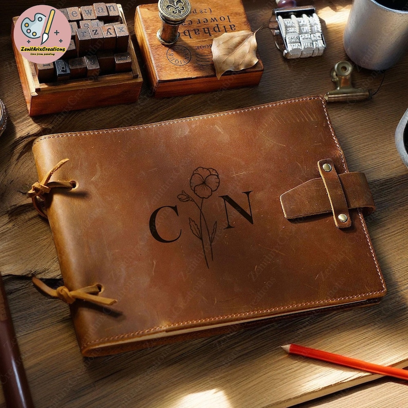 May include: A brown leather journal with a floral design and the initials "CN" embossed on the front cover. The journal has a strap closure and is lying on a wooden surface.