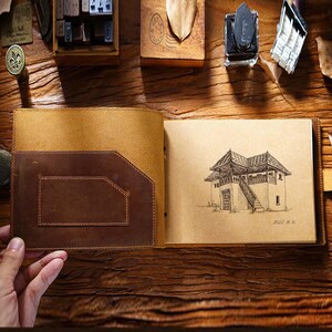 May include: A brown leather journal with a tan suede cover and a flap closure. The journal is open to a page with a pen and ink drawing of a house with a staircase. The drawing is dated "2022 8.6".