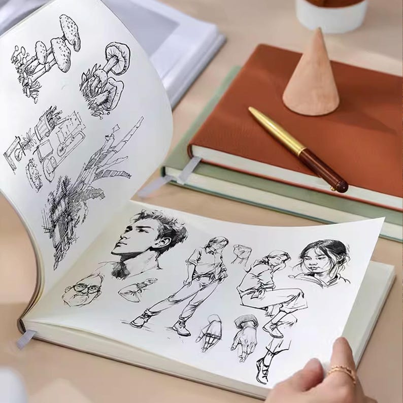 May include: An open sketchbook displaying detailed black and white sketches of mushrooms, machinery, and human figures. A brown and gold pen rests on a closed brown notebook. A small wooden cone sits nearby.