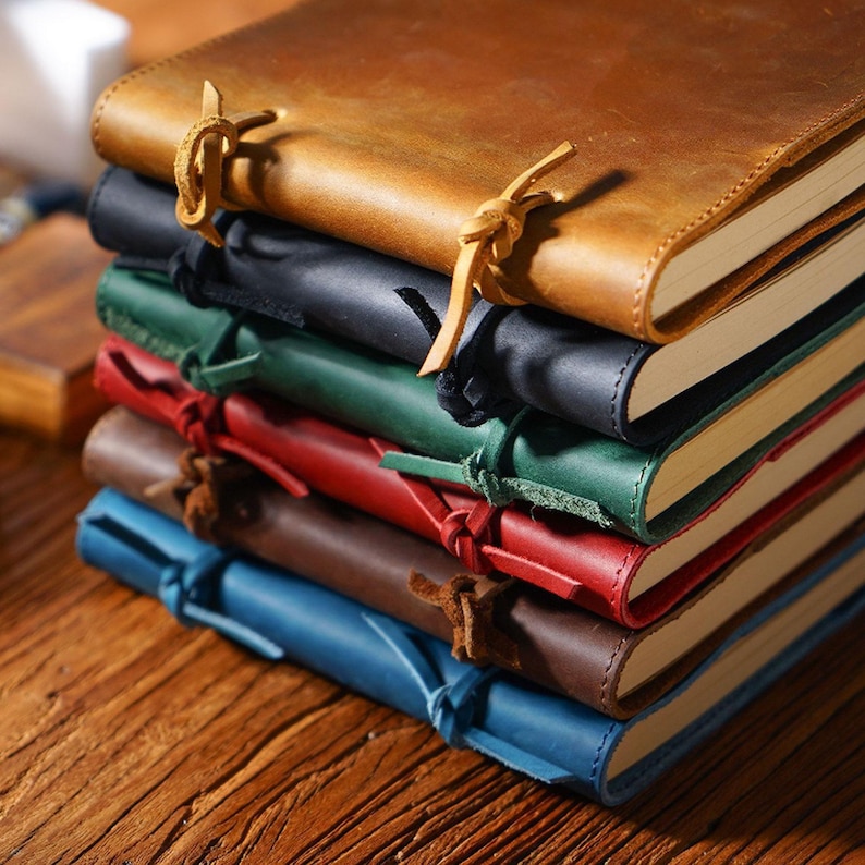 May include: A stack of leather-bound journals with different colored covers. The top journal is brown and has a leather strap closure. The journals are stacked on a wooden surface.